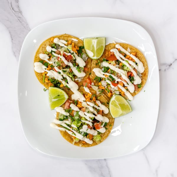 Three Olivo Taco tacos with meat, pico de gallo, crema, and lime wedges on a white plate.