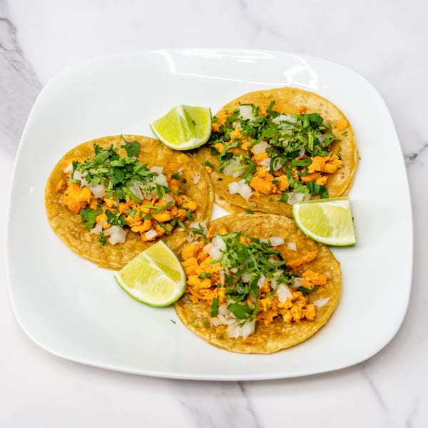 Three Olivo Taco al pastor tacos with cilantro, onions, and lime wedges on a white plate.