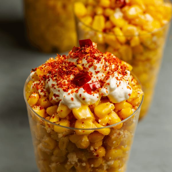 Two clear cups of Mexican esquites, topped with white cream and red chili flakes.